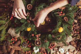 Hands making a Christmas wreath with foliage, cinnamon, orange and pine cones.