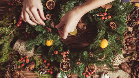 Hands making a Christmas wreath with foliage, cinnamon, orange and pine cones.