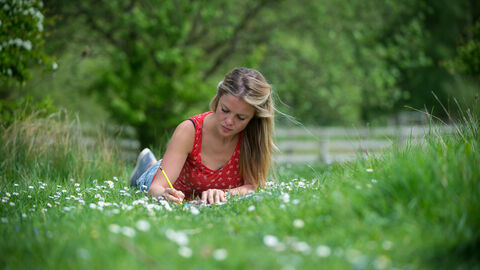A woman lying on grass covered with daisies, writing. There are trees and bushes visible around her