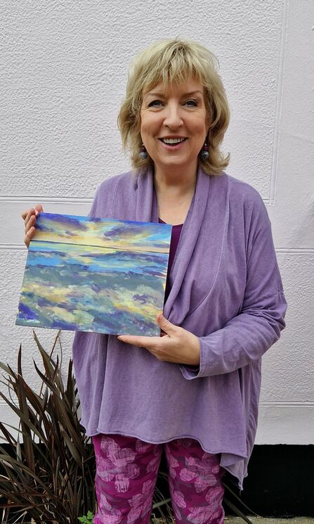 A lady dressed in purple holds her ocean sunset artwork.