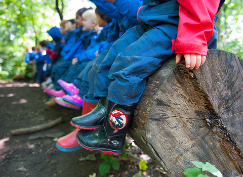 A line of children sat on a tree trunk wearing wellies, spiderman ones at the forefront